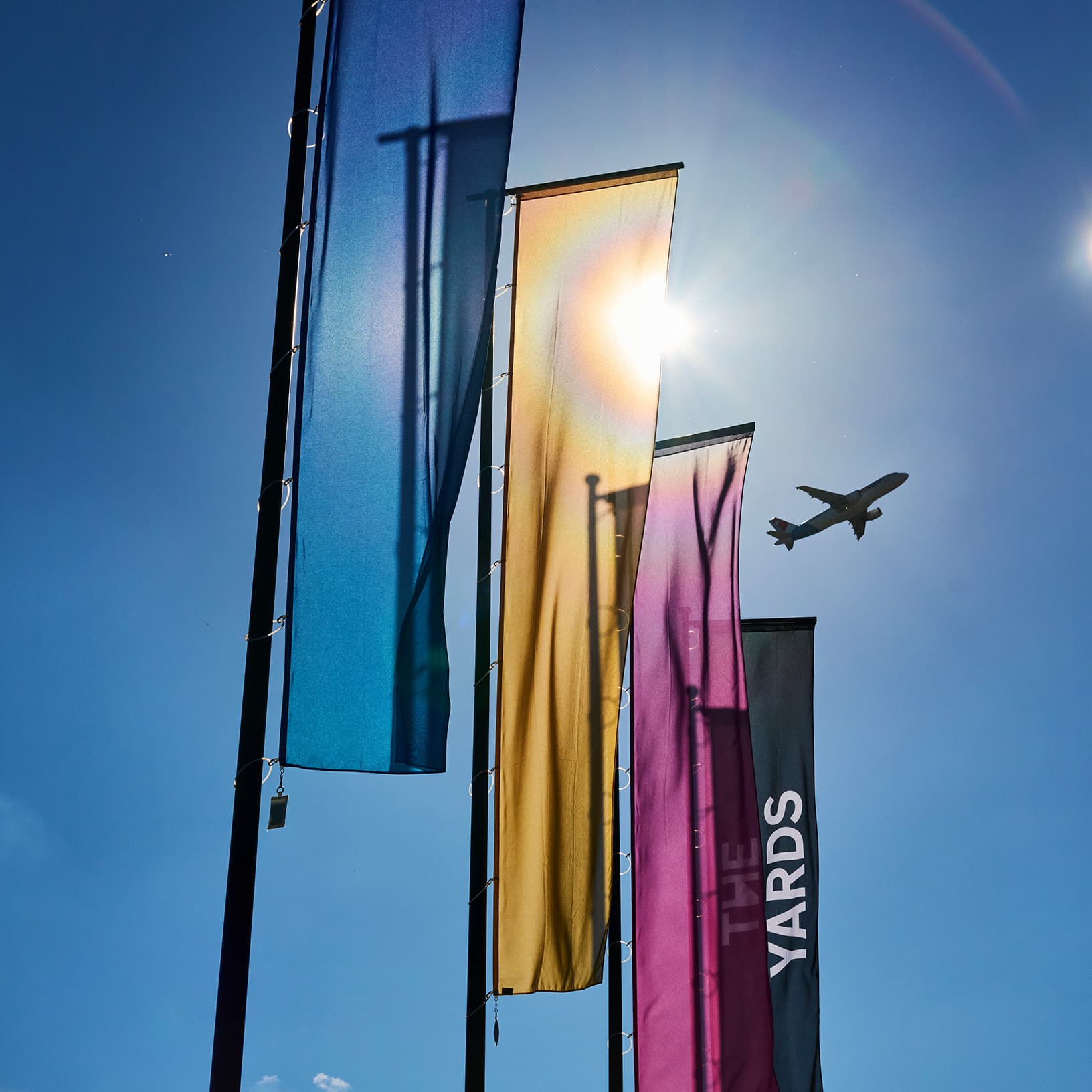  Farbige The Yards Fahnen vor blauem Himmel, dahinter ein vorbeifliegendes Flugzeug in der Flughafenregion Zürich.