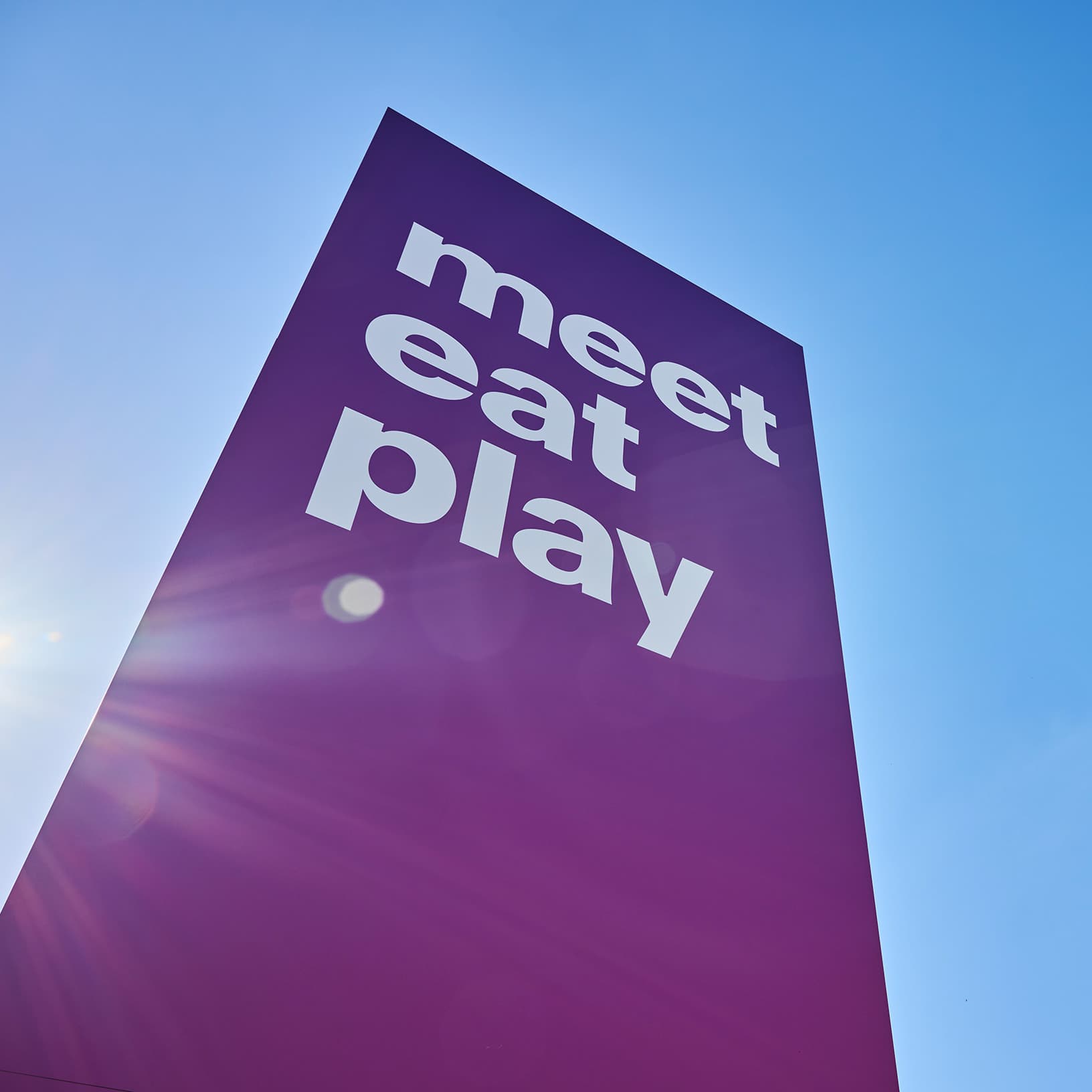 Violette The Yards Stele mit dem weissen Schriftzug «meet eat play» vor blauem Himmel und Sonnenlicht.