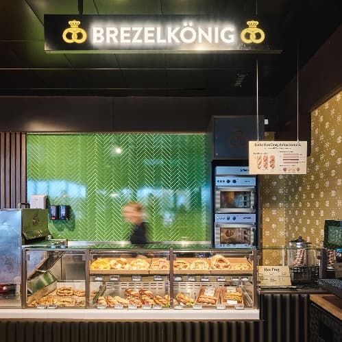 Brezelkönig bei The Yards: Verkaufstheke mit Brezelauslage, grün gefliester Wand und beleuchtetem Logo.
