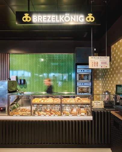 Frontalansicht der Brezelkönig-Verkaufstheke mit Backwarenauslage, grünen Fliesen und beleuchtetem Logo.
