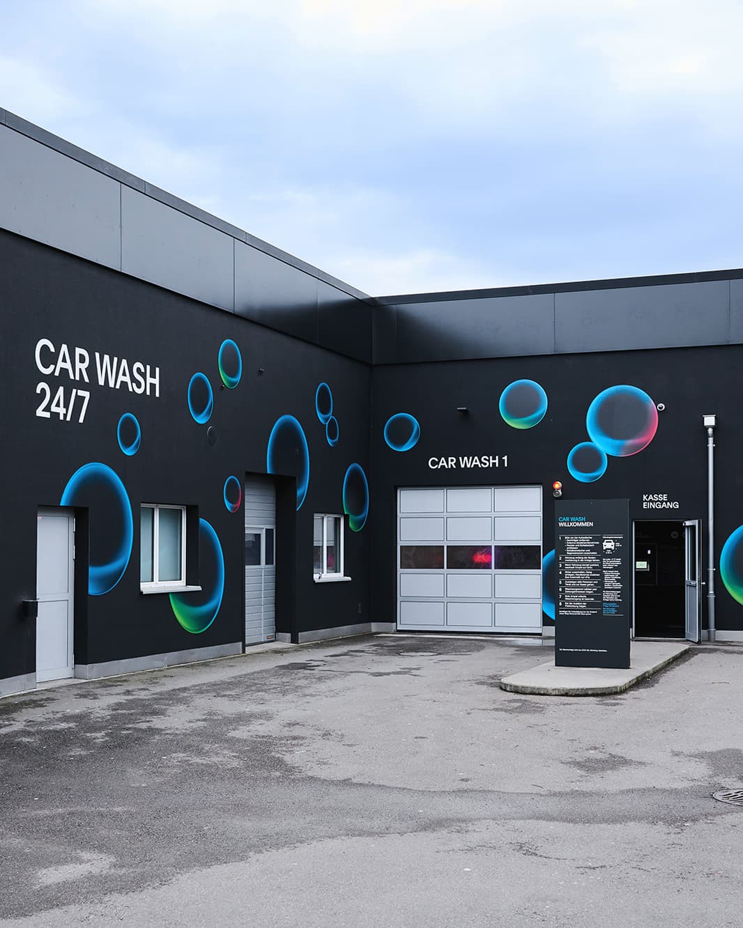 Einfahrt zur Car Wash 24/7 mit Waschhalle, Kassenbereich und blauen Kreismotiven bei The Yards in Rümlang.
