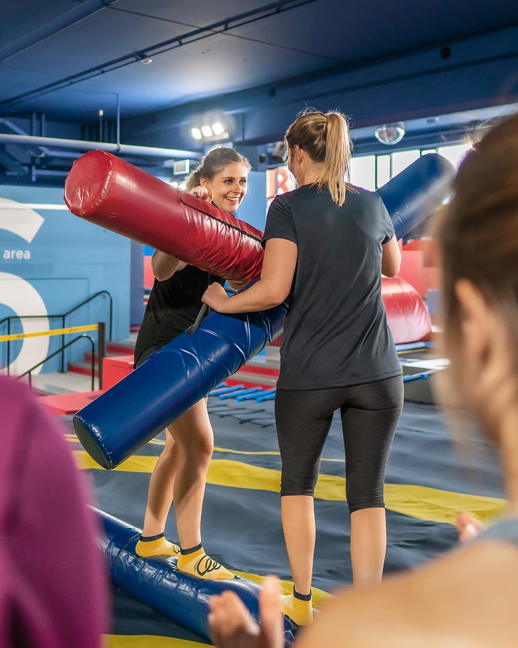 Zwei Frauen spielen Battle Beam mit gepolsterten Stangen über einem Balancierbalken im Flip Lab bei The Yards.