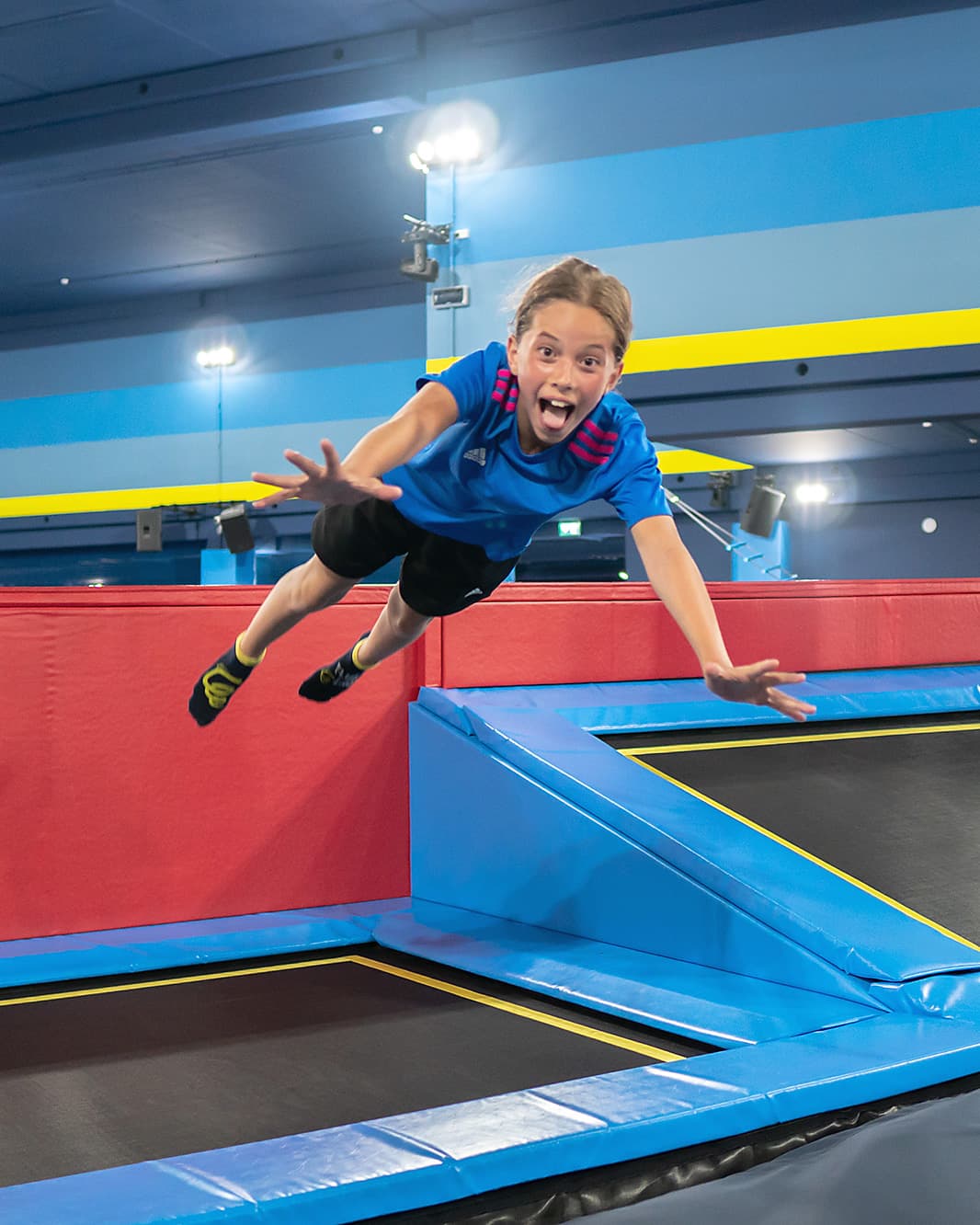 Junge in blauem T-Shirt fliegt mit ausgestreckten Armen über ein Trampolin im Flip Lab bei The Yards.