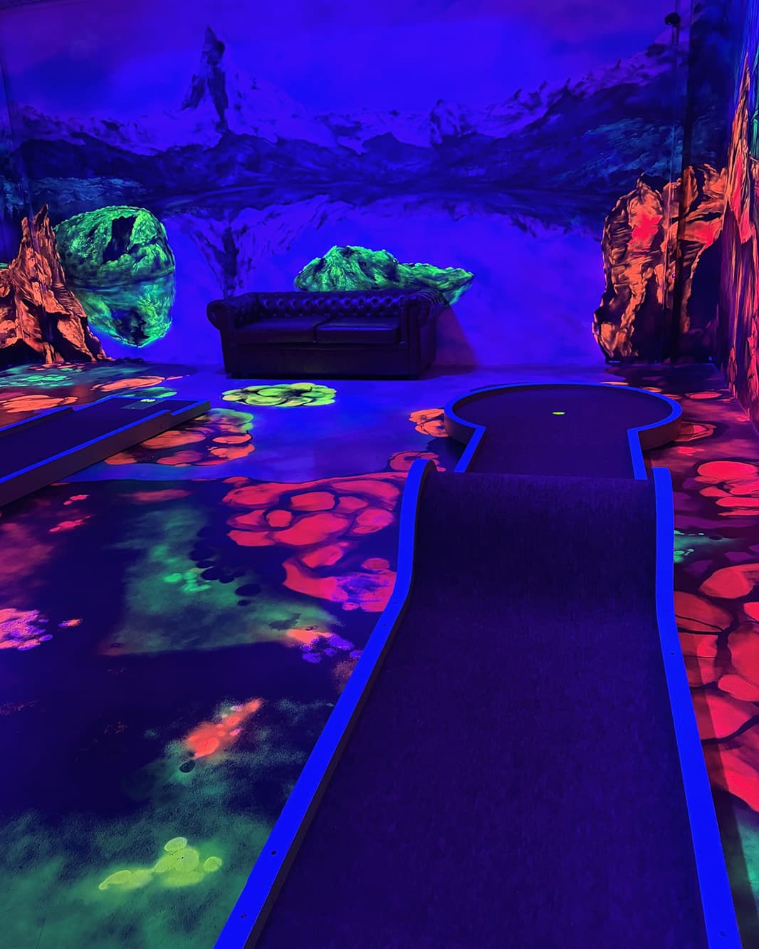 Schwarzlicht-Minigolfbahn vor einer blau-violetten Fantasielandschaft mit leuchtenden Felsen bei Galaxy Golf.