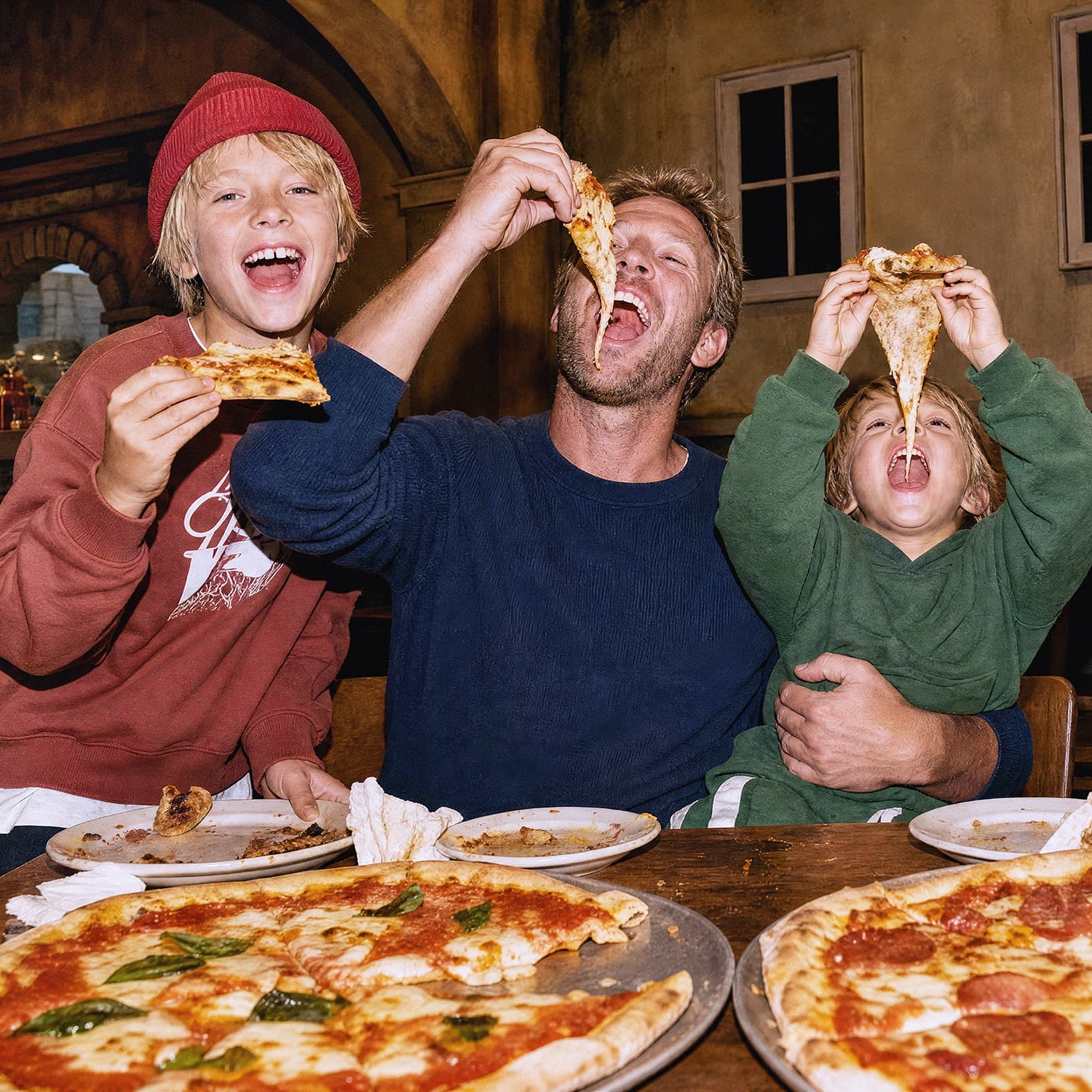 Ein Vater und zwei Kinder essen lachend Pizza im Restaurant Piazza Meraviglia bei The Yards – ein Motiv für Familienausflüge und gemeinsames Essen.