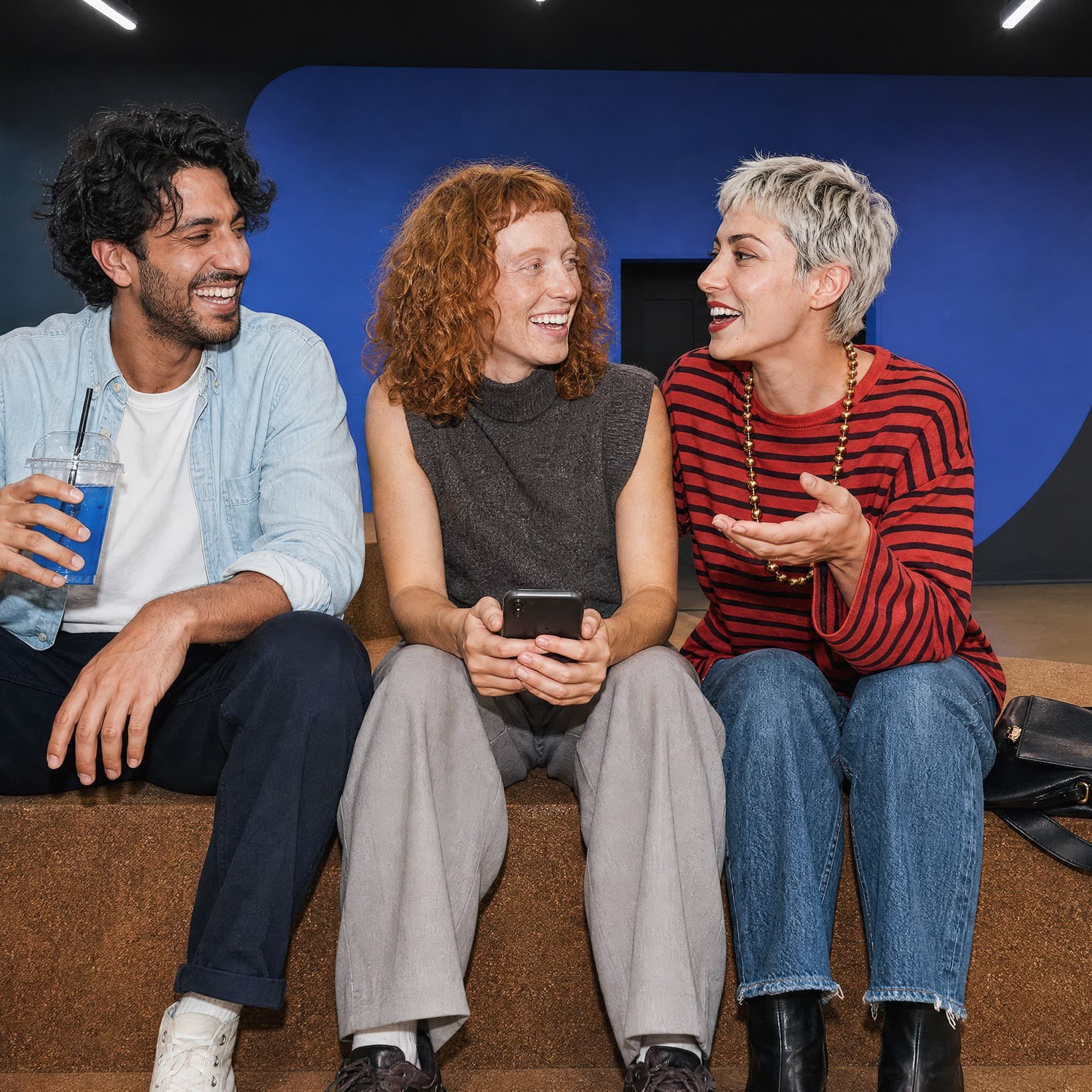 Drei Erwachsene sitzen vor einer blauen Wand auf den Sitzstufen in The Yards und unterhalten sich lachend – ein Motiv für Teamevents.