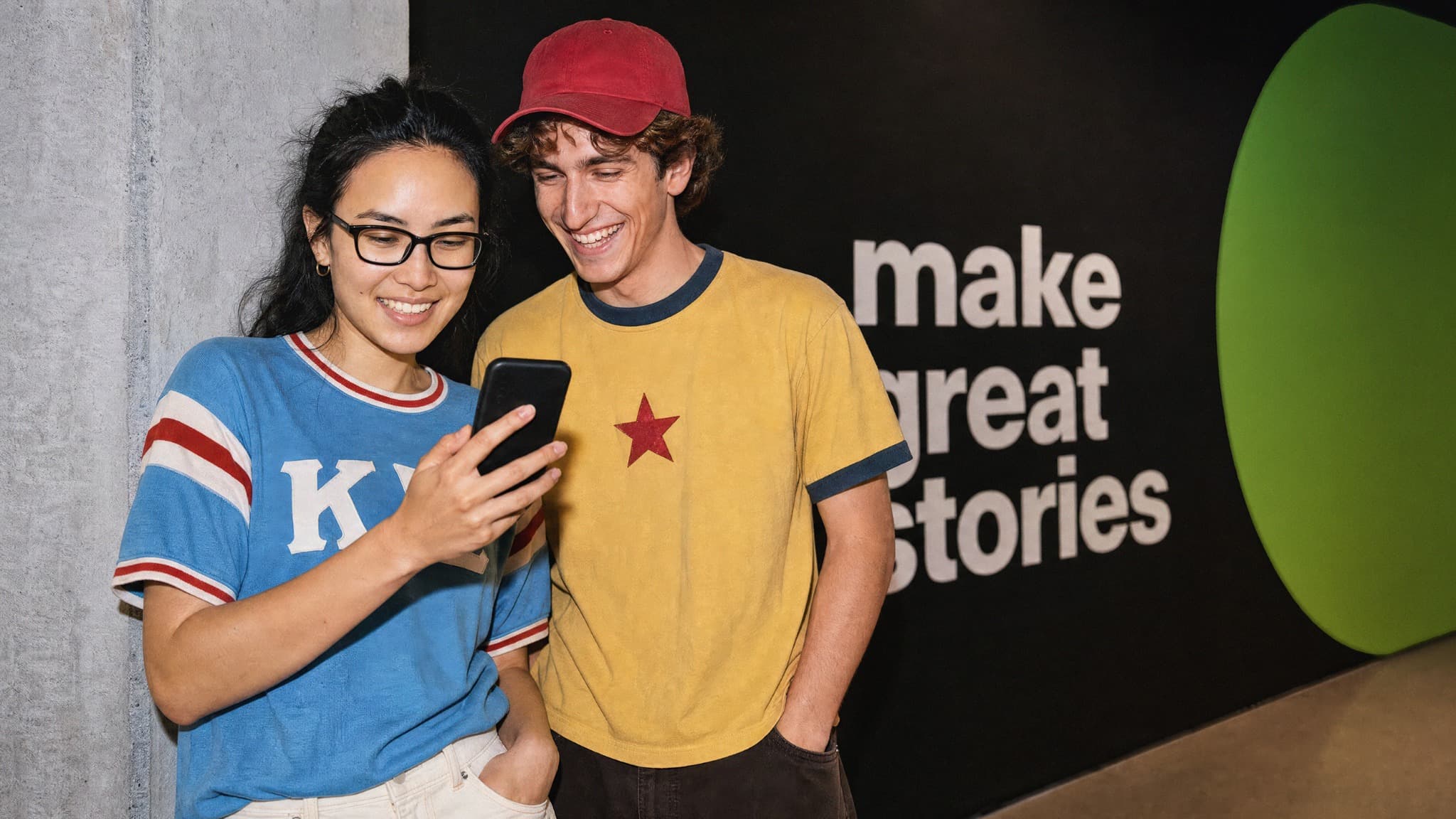 Zwei junge Erwachsene betrachten lächelnd ein Smartphone vor dem Schriftzug «make great stories» bei The Yards.