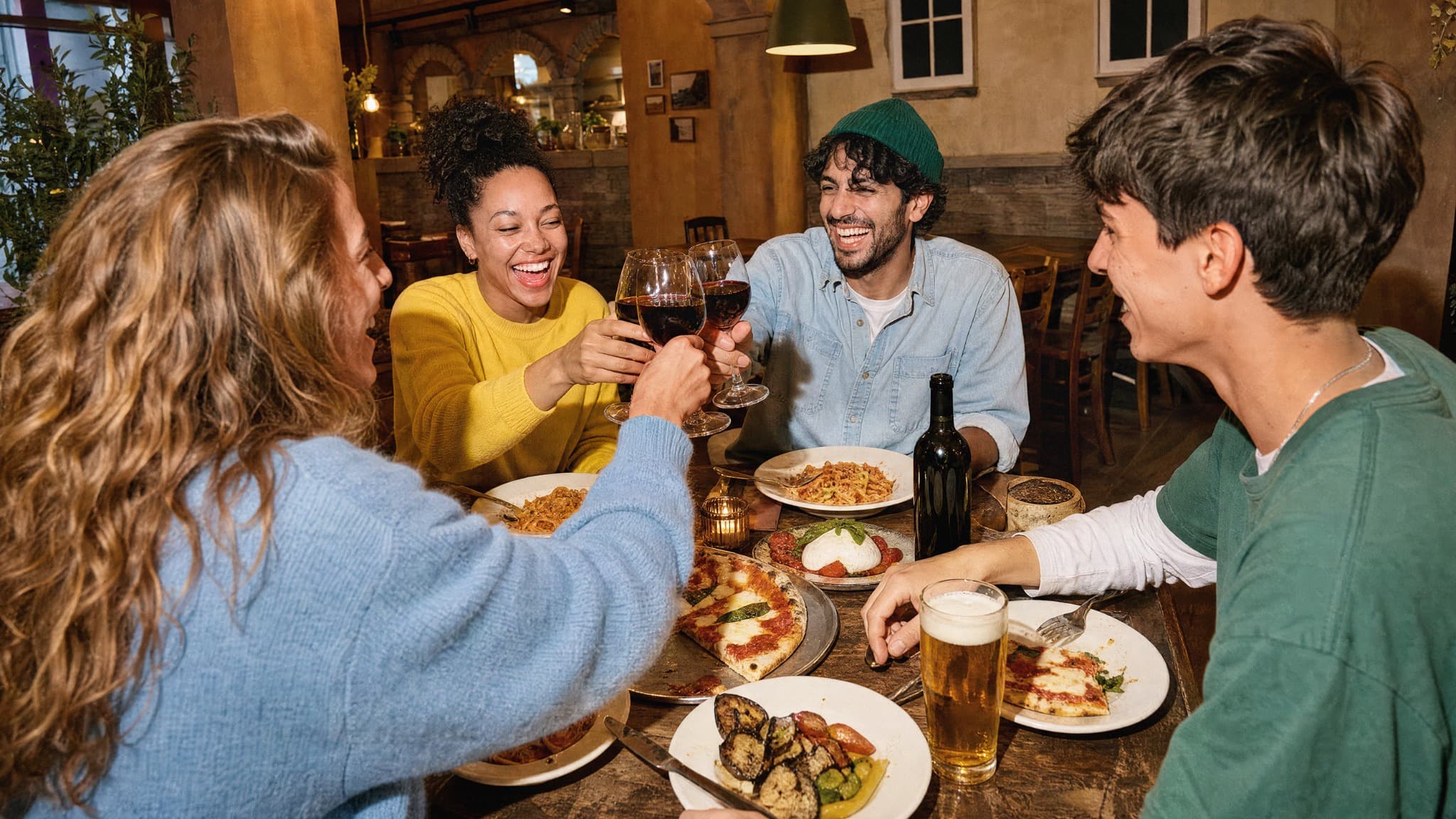 Vier Erwachsene stossen am gedeckten Tisch mit Weingläsern an und geniessen Pizza und Pasta im Restaurant Piazza Meraviglia bei The Yards.