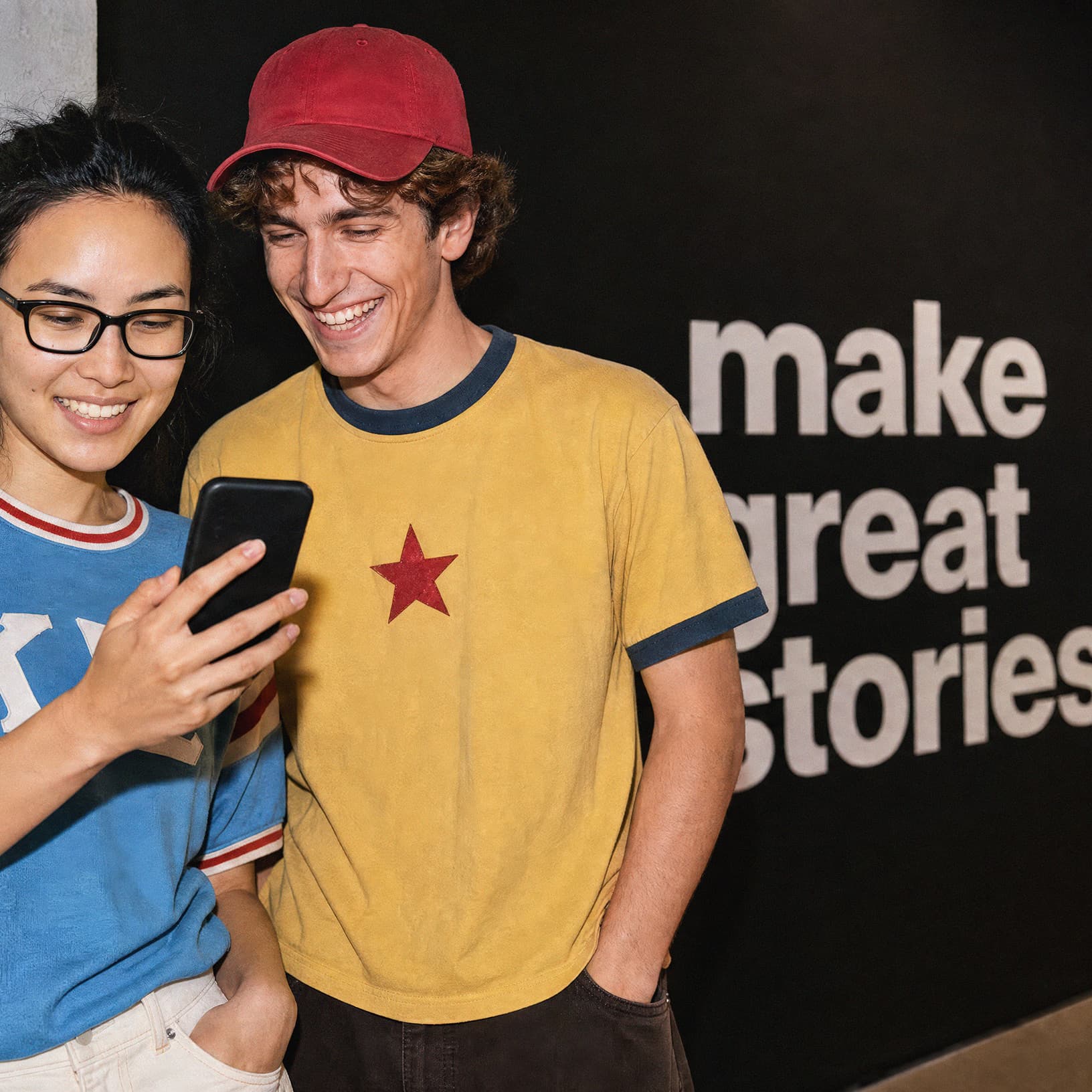 Zwei junge Erwachsene schauen lächelnd auf ein Smartphone vor dem Schriftzug «make great stories» in The Yards.