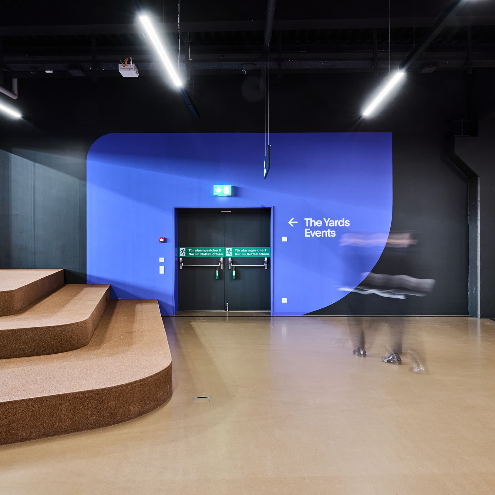 Blau-violett gestaltete Wand mit Wegweiser zu The Yards Events, daneben Sitzstufen und vorbeigehende Menschen.