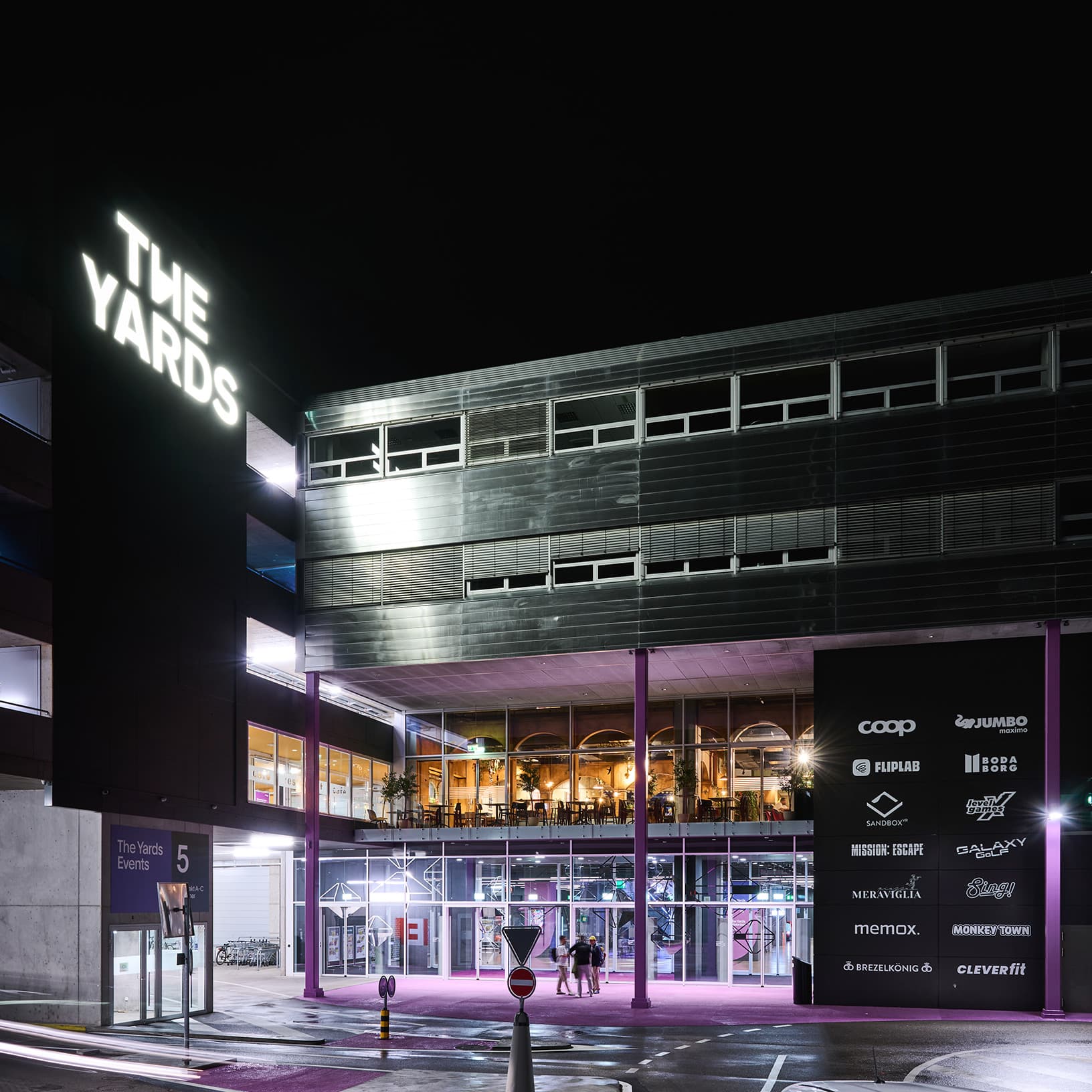 The Yards in Rümlang bei Nacht mit beleuchtetem Logo, Haupteingang und Übersicht der Erlebnis-, Gastronomie- und Einkaufsangebote.