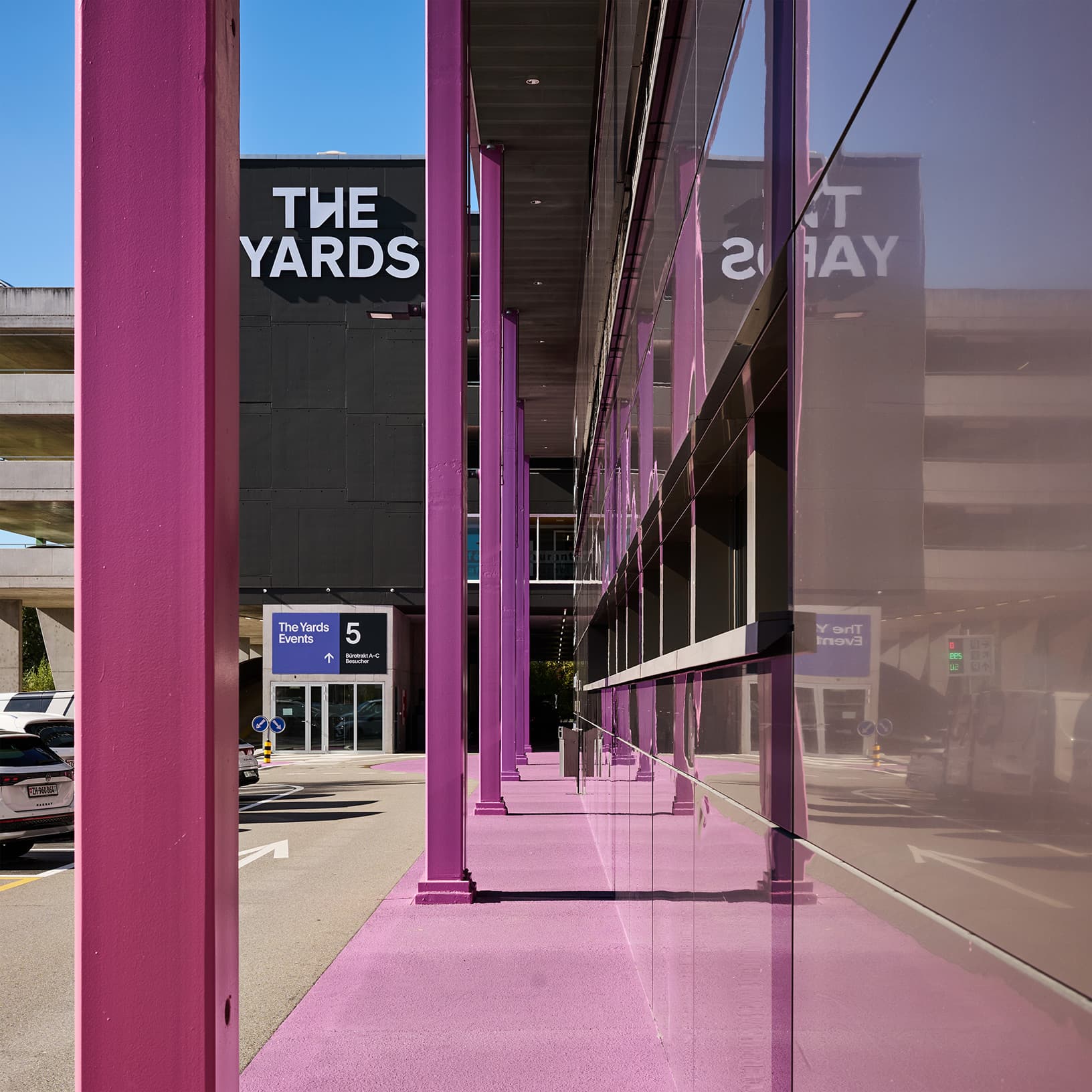 Violette Säulen entlang der spiegelnden Glasfassade von The Yards in Rümlang, mit dem Markenlogo im Hintergrund.