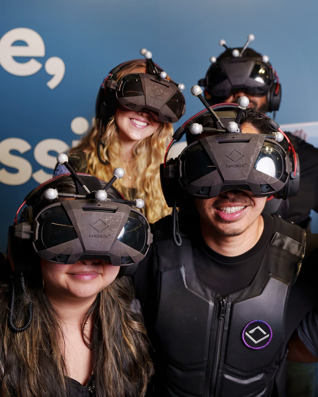 Vier Personen mit VR-Headsets und Tracking-Ausrüstung stehen lächelnd als Gruppe bei Sandbox VR.
