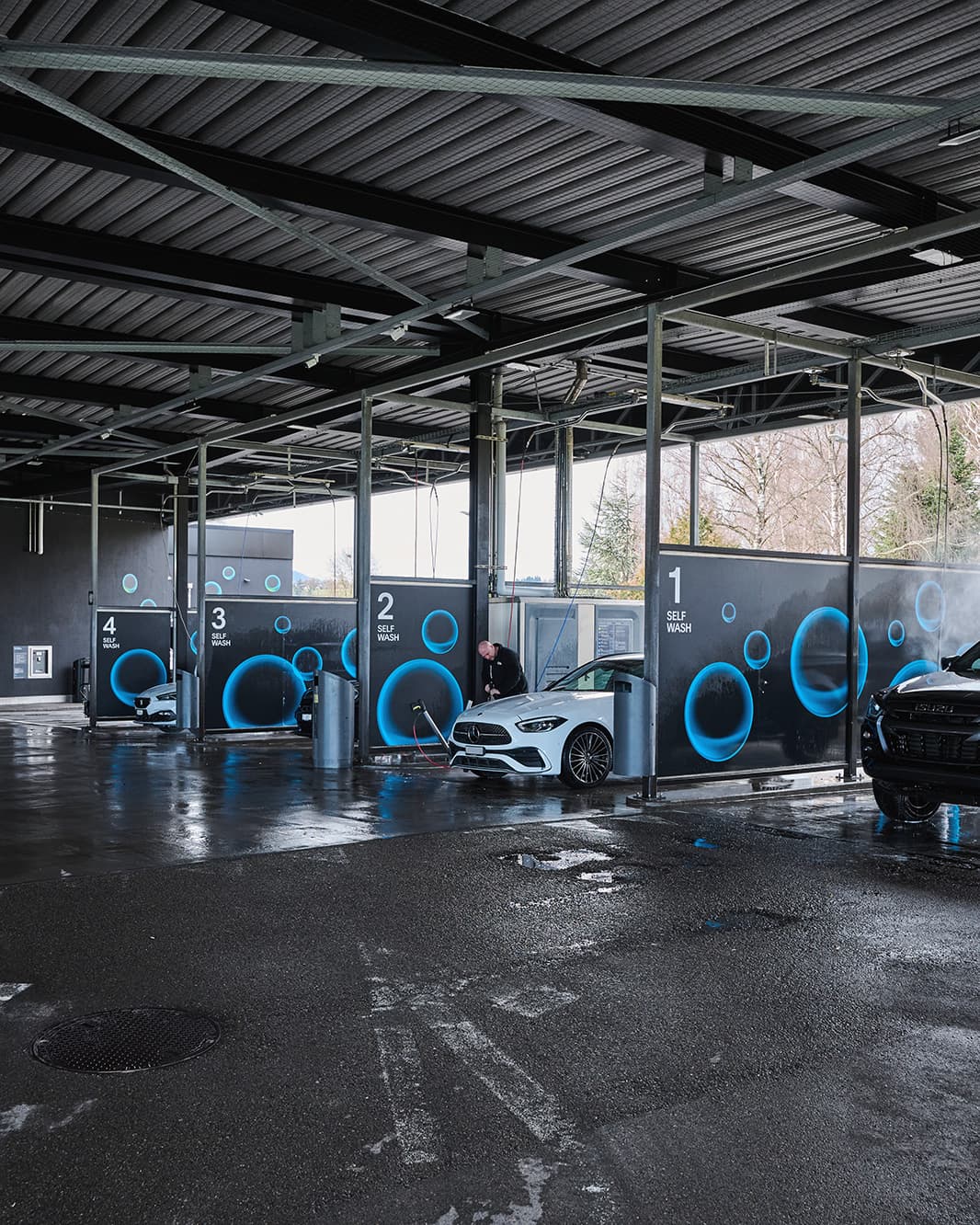 Vier überdachte Self-Wash-Waschplätze mit Autos und blauen Kreisgrafiken bei The Yards in Rümlang.